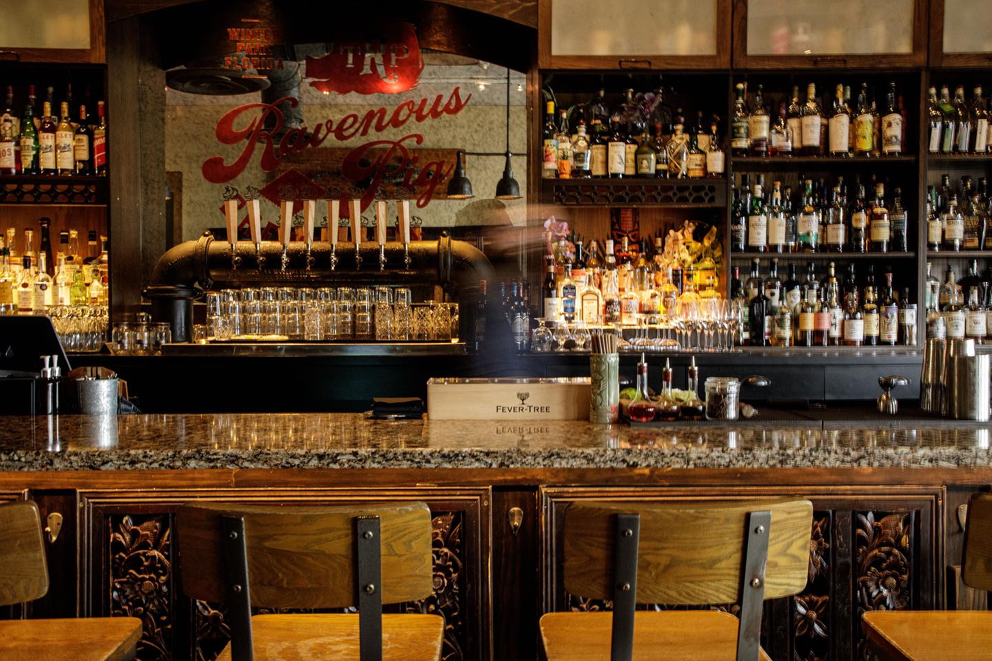 A cozy bar lined with bottles and warm lighting sets the mood for a relaxed evening drink. The wooden counter and vintage details give the space a rich, welcoming feel. It looks like the kind of bar where you unwind with great cocktails and good company.