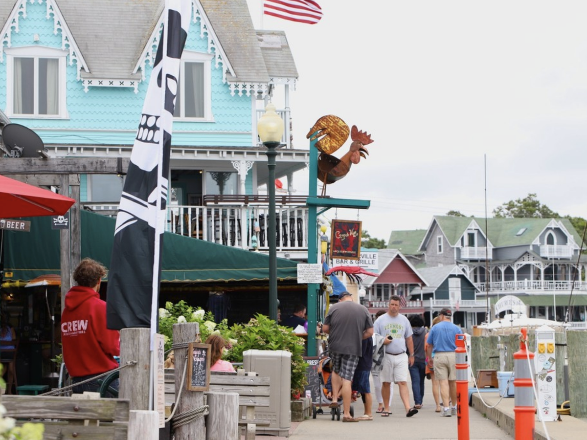 Busy waterfront boardwalk lined with colorful Victorian-style buildings, restaurants, and shops, with people walking along the marina under an overcast sky