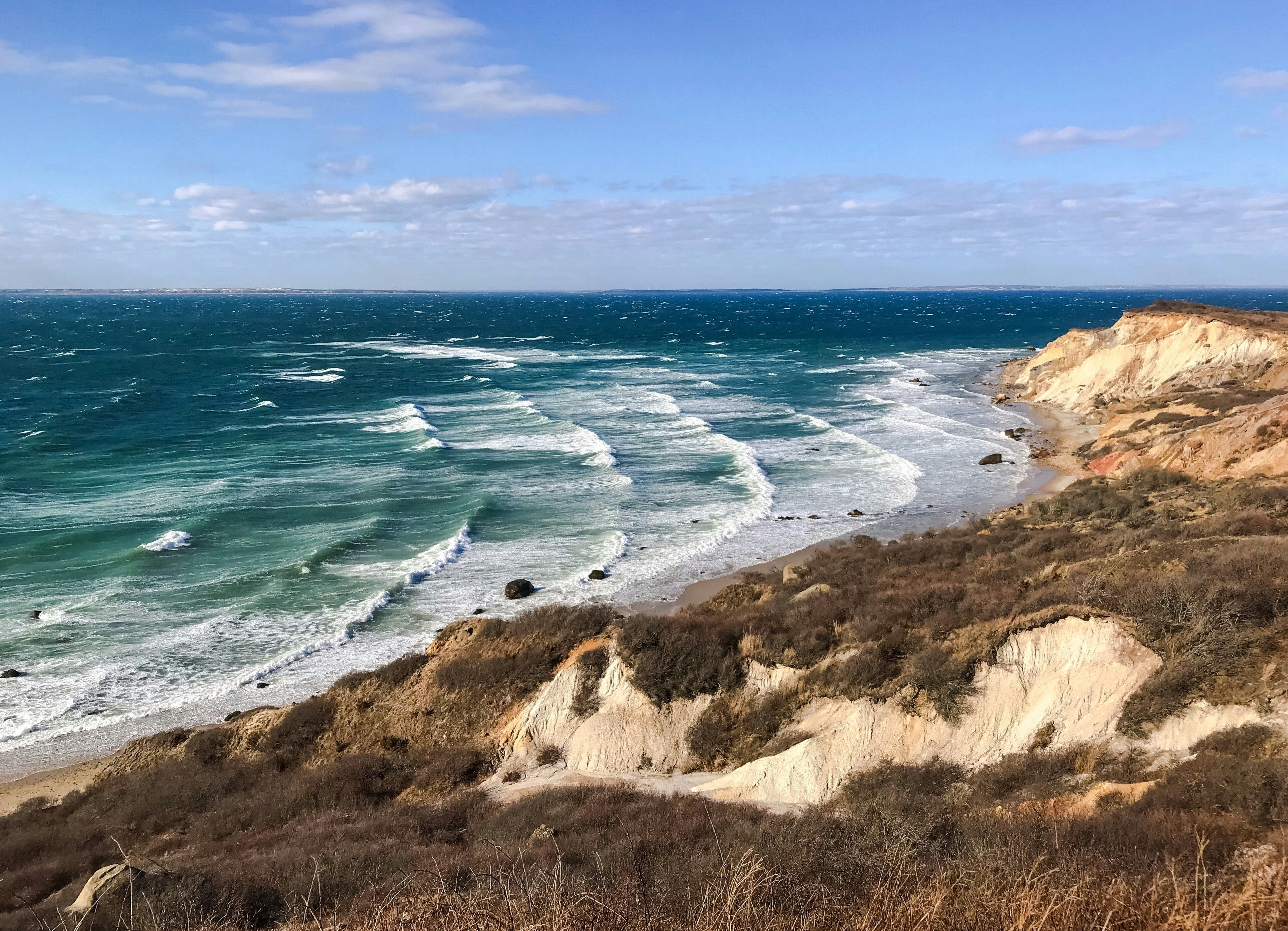 Waves crash against the rugged, windswept cliffs of Martha’s Vineyard’s Aquinnah shoreline under a bright blue sky.