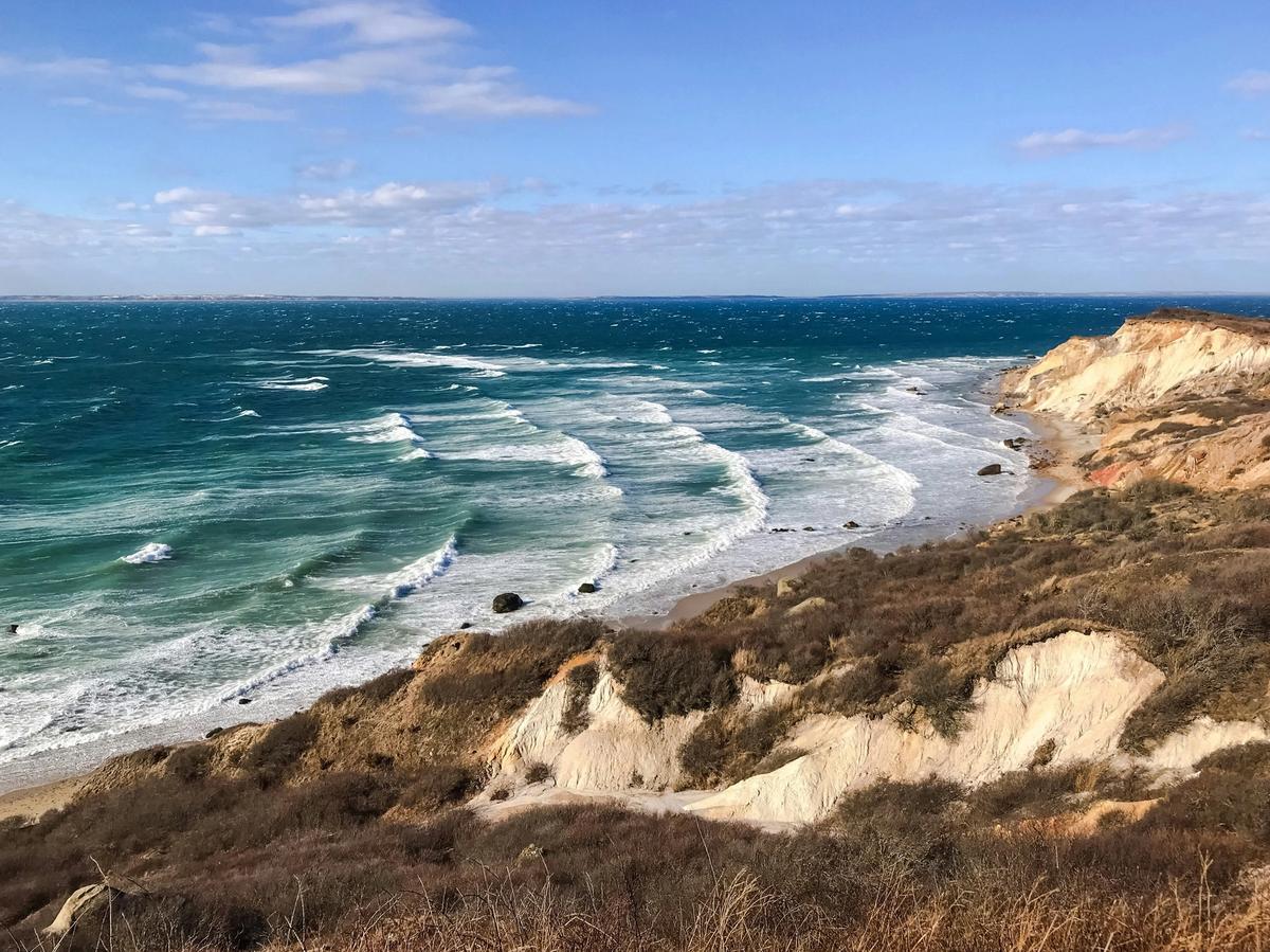 Waves crash against the rugged, windswept cliffs of Martha’s Vineyard’s Aquinnah shoreline under a bright blue sky.
