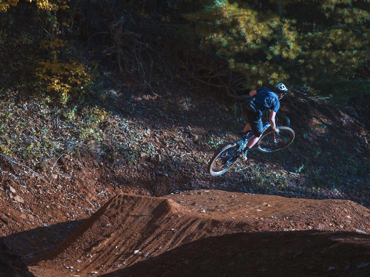 A mountain biker catches air off a dirt jump on a forested trail, surrounded by autumn foliage and dappled sunlight.