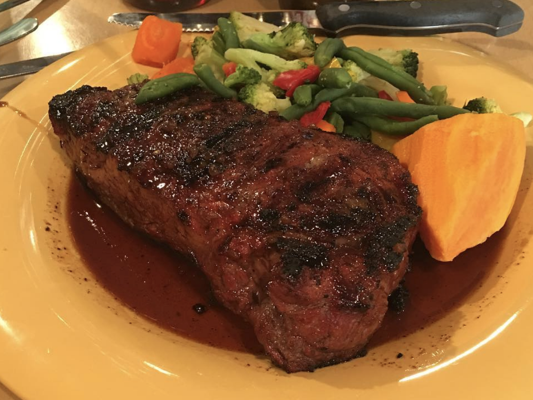 A grilled steak served with a side of steamed mixed vegetables and a large slice of sweet potato on a yellow plate.