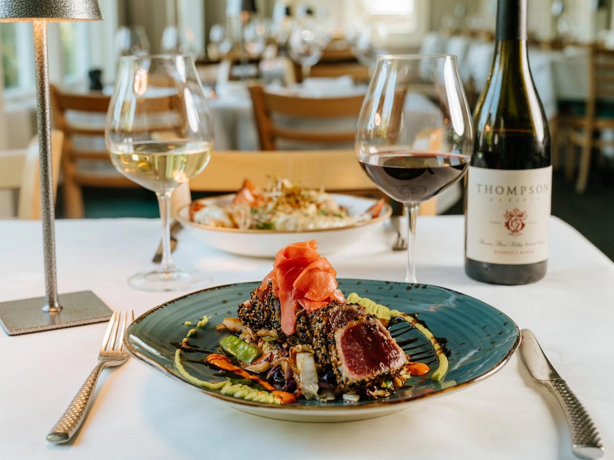 A seared tuna dish sits on a stylish plate, surrounded by colorful vegetables, sauces, and glasses of red and white wine. The warm lighting and clean table setting create a classy coastal dining vibe. It feels fresh, modern, and perfect for a special night out.