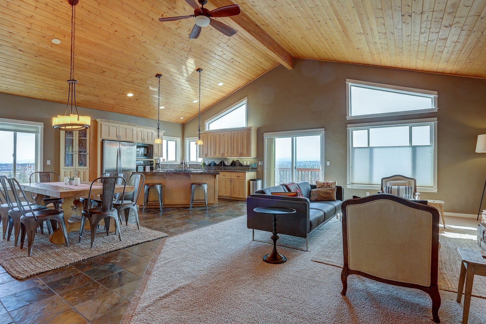 A vaulted knotty-pine ceiling with a wood ceiling fan tops this open living and dining space, where a charcoal sofa and upholstered chairs face windows that frame a distant tree-lined view, alongside a reclaimed-wood dining table ringed with metal chairs. Slate tile and a granite-topped kitchen island round out the great room, capturing the elevated indoor-outdoor feel of a hilltop vacation home in Central Oregon.