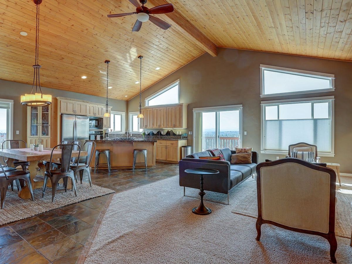A vaulted knotty-pine ceiling with a wood ceiling fan tops this open living and dining space, where a charcoal sofa and upholstered chairs face windows that frame a distant tree-lined view, alongside a reclaimed-wood dining table ringed with metal chairs. Slate tile and a granite-topped kitchen island round out the great room, capturing the elevated indoor-outdoor feel of a hilltop vacation home in Central Oregon.