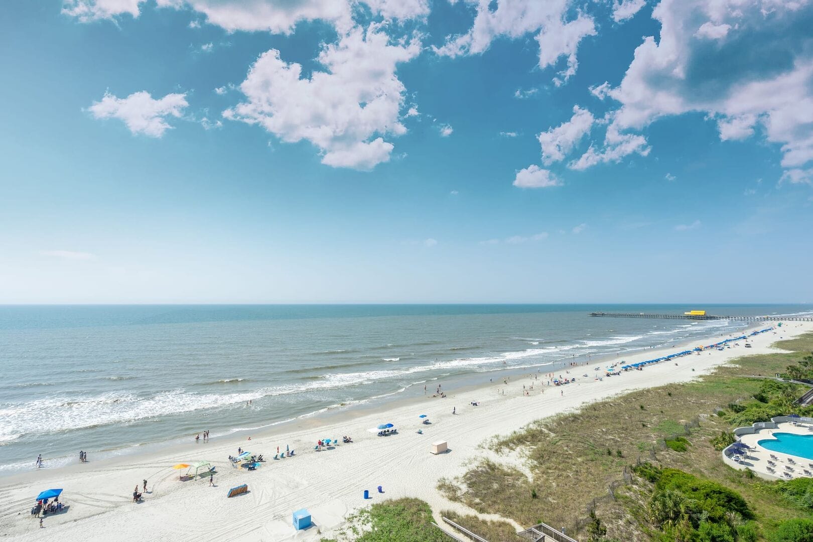 A wide sandy shoreline stretches along Myrtle Beach, South Carolina, with gentle waves rolling in under a bright sky. Beachgoers relax under umbrellas while the pier extends into the Atlantic Ocean. Myrtle Beach is known for its long coastline and fun summer atmosphere.