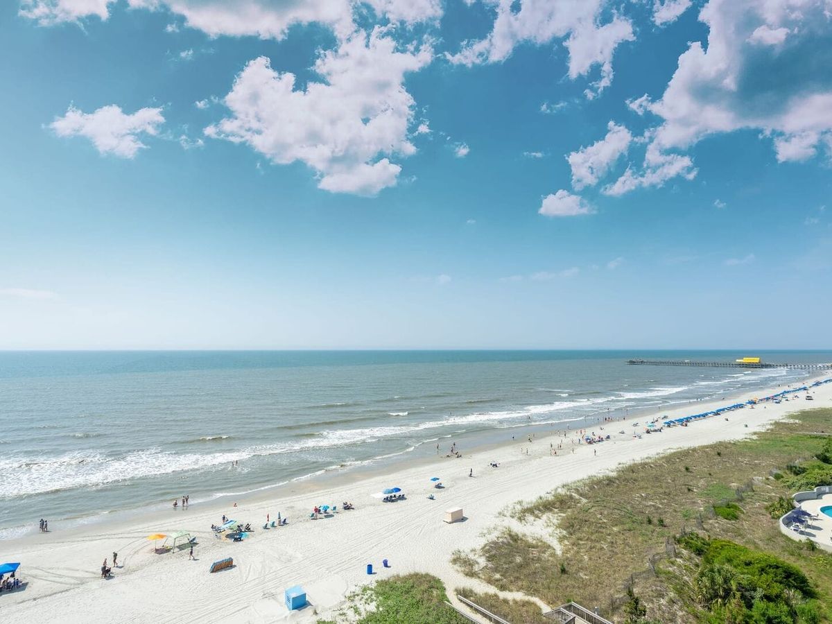 A wide sandy shoreline stretches along Myrtle Beach, South Carolina, with gentle waves rolling in under a bright sky. Beachgoers relax under umbrellas while the pier extends into the Atlantic Ocean. Myrtle Beach is known for its long coastline and fun summer atmosphere.