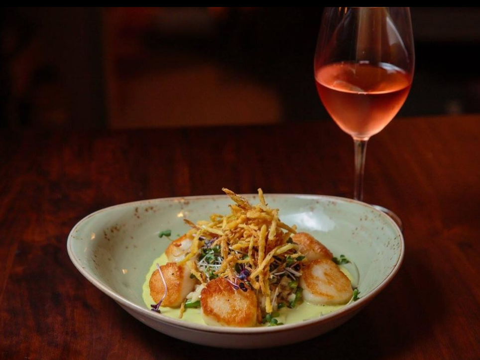 A beautifully plated dish of seared scallops with crispy garnish sits beside a glass of rosé on a wooden table in warm, ambient lighting