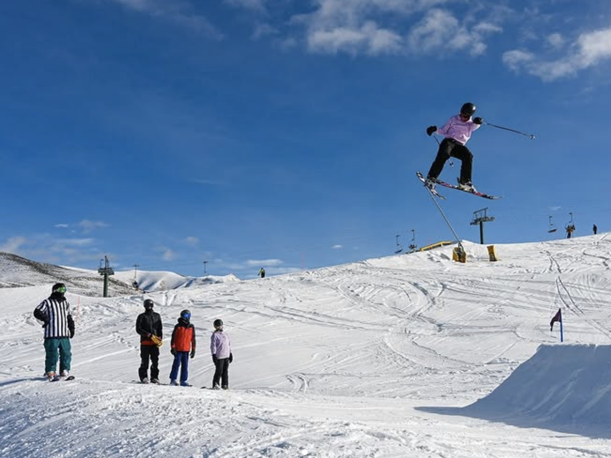 A skier launches off a snow ramp mid-air while a group of people watches nearby on a sunny ski slope with chairlifts in the background.