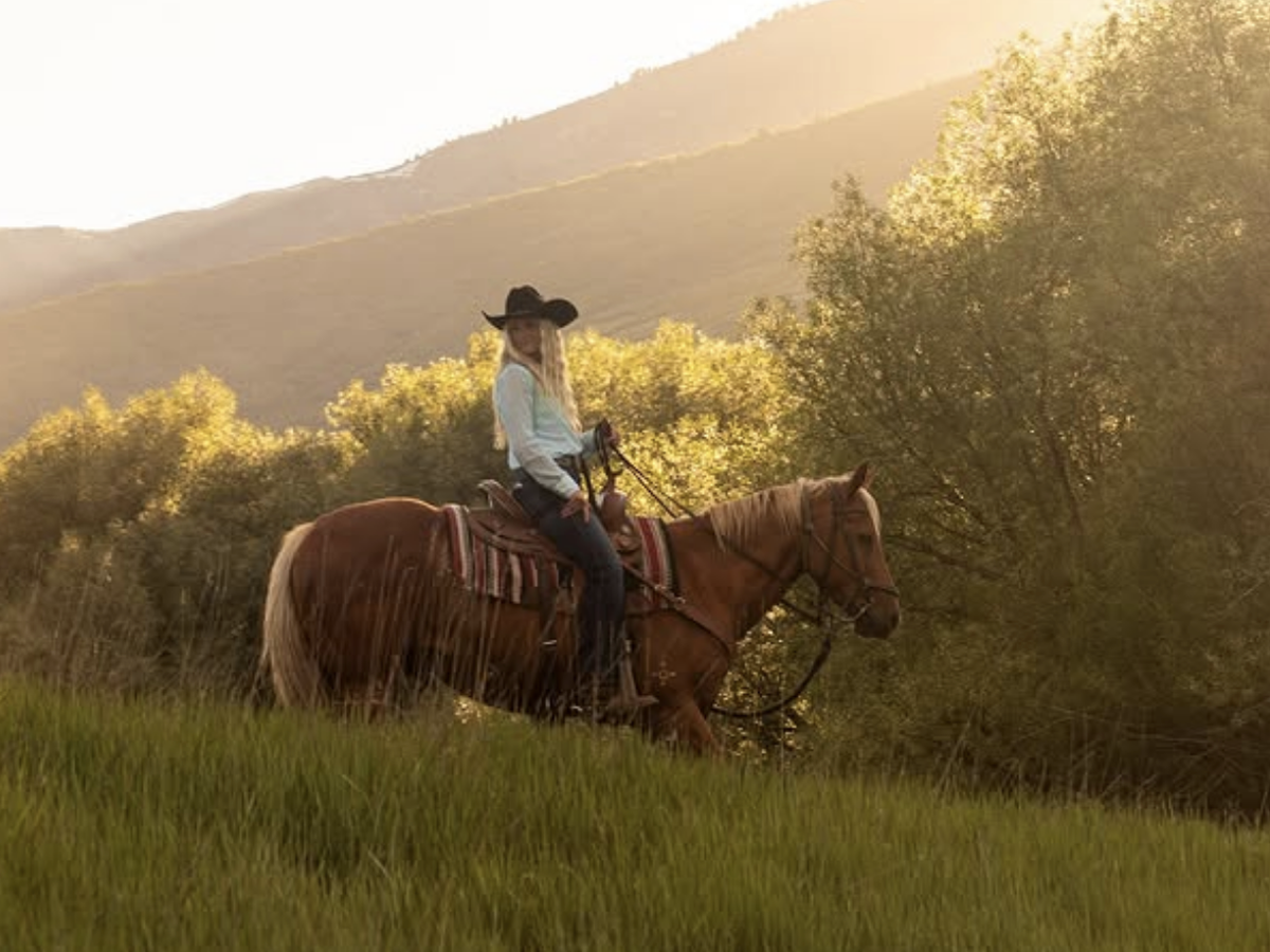 Cowgirl with long blonde hair rides a brown horse through a grassy field at golden hour, with sunlight streaming through trees and mountains in the background.
