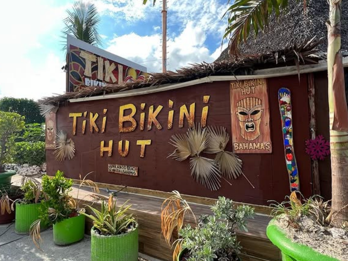 A tropical-themed bar called “Tiki Bikini Hut” in the Bahamas, with a thatched roof, tiki masks, colorful signage, and vibrant green planters bursting with island foliage.