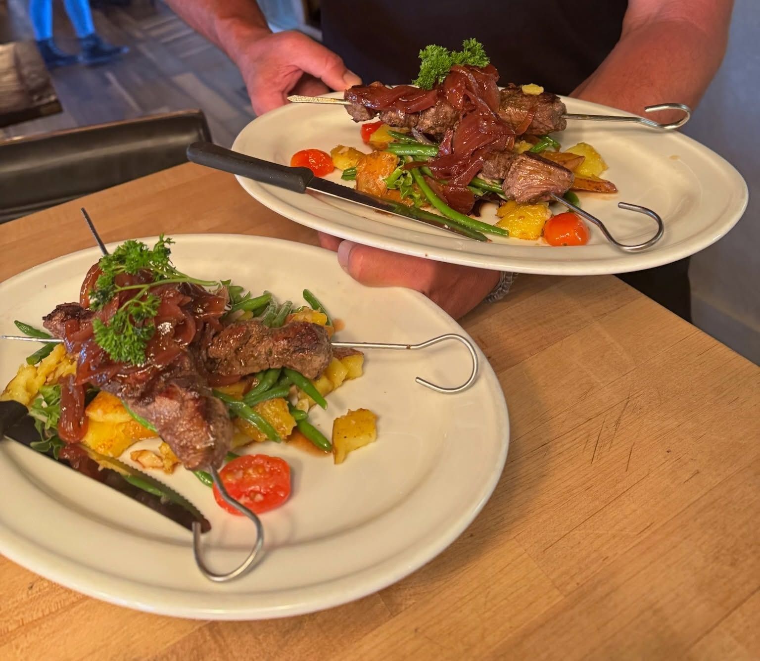 A server carries two plates of grilled beef skewers topped with caramelized red onions and fresh parsley, set over roasted potatoes, green beans, and cherry tomatoes. The plates rest on a butcher block counter in a bustling kitchen. 