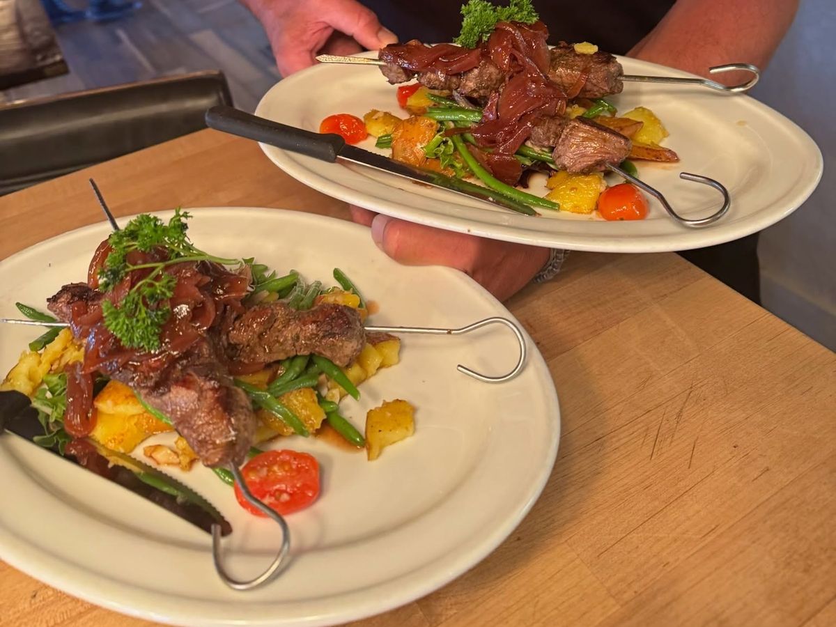 A server carries two plates of grilled beef skewers topped with caramelized red onions and fresh parsley, set over roasted potatoes, green beans, and cherry tomatoes. The plates rest on a butcher block counter in a bustling kitchen.