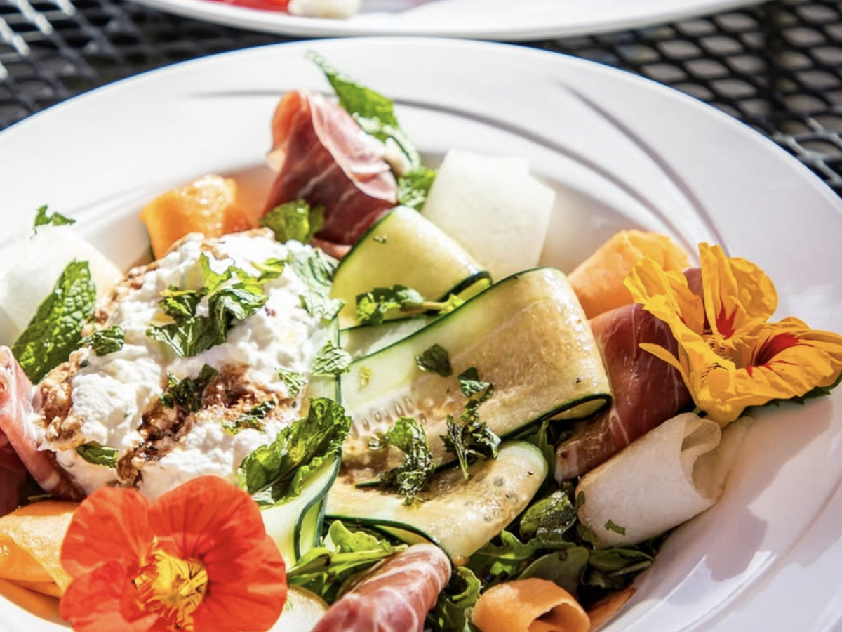 Fresh summer salad with prosciutto, zucchini ribbons, melon, ricotta, mint, and edible flowers served on a white plate.