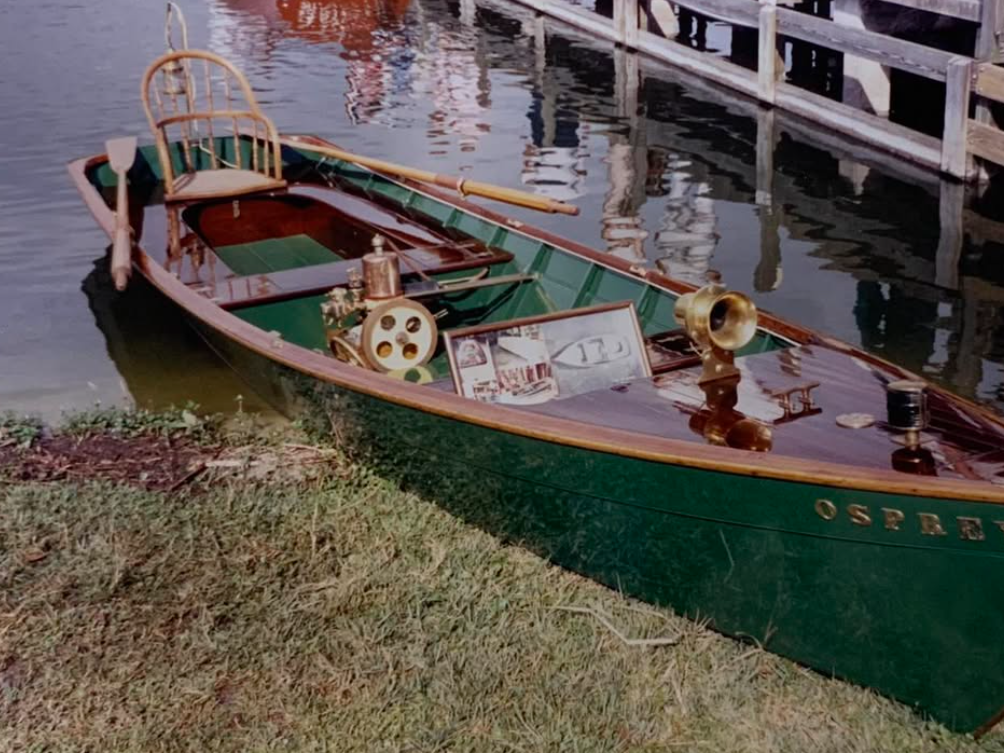 A vintage green wooden boat named "Osprey" is docked beside the shore, featuring polished brass fittings, a wooden chair, and classic nautical instruments.