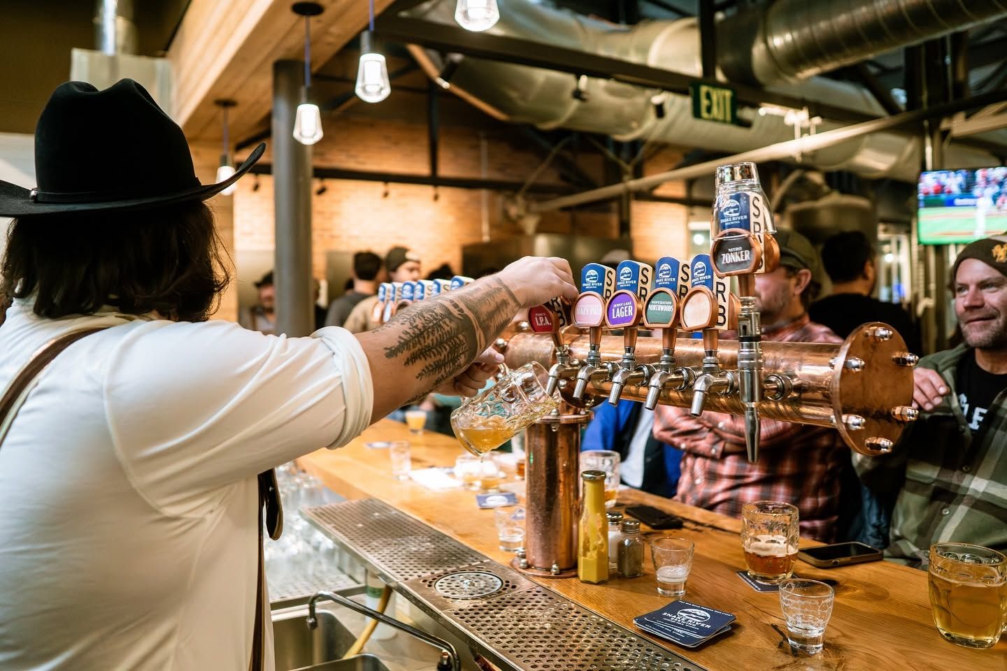  A bartender in a black cowboy hat pours a fresh beer from a gleaming copper tap tower lined with Snake River Brewing tap handles. Patrons gather along the wooden bar in the lively, industrial-style brewpub with exposed beams and ductwork overhead