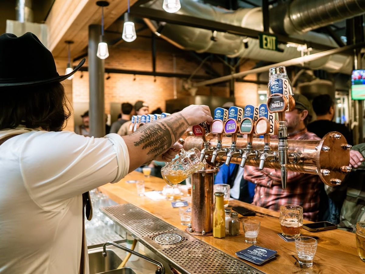 A bartender in a black cowboy hat pours a fresh beer from a gleaming copper tap tower lined with Snake River Brewing tap handles. Patrons gather along the wooden bar in the lively, industrial-style brewpub with exposed beams and ductwork overhead