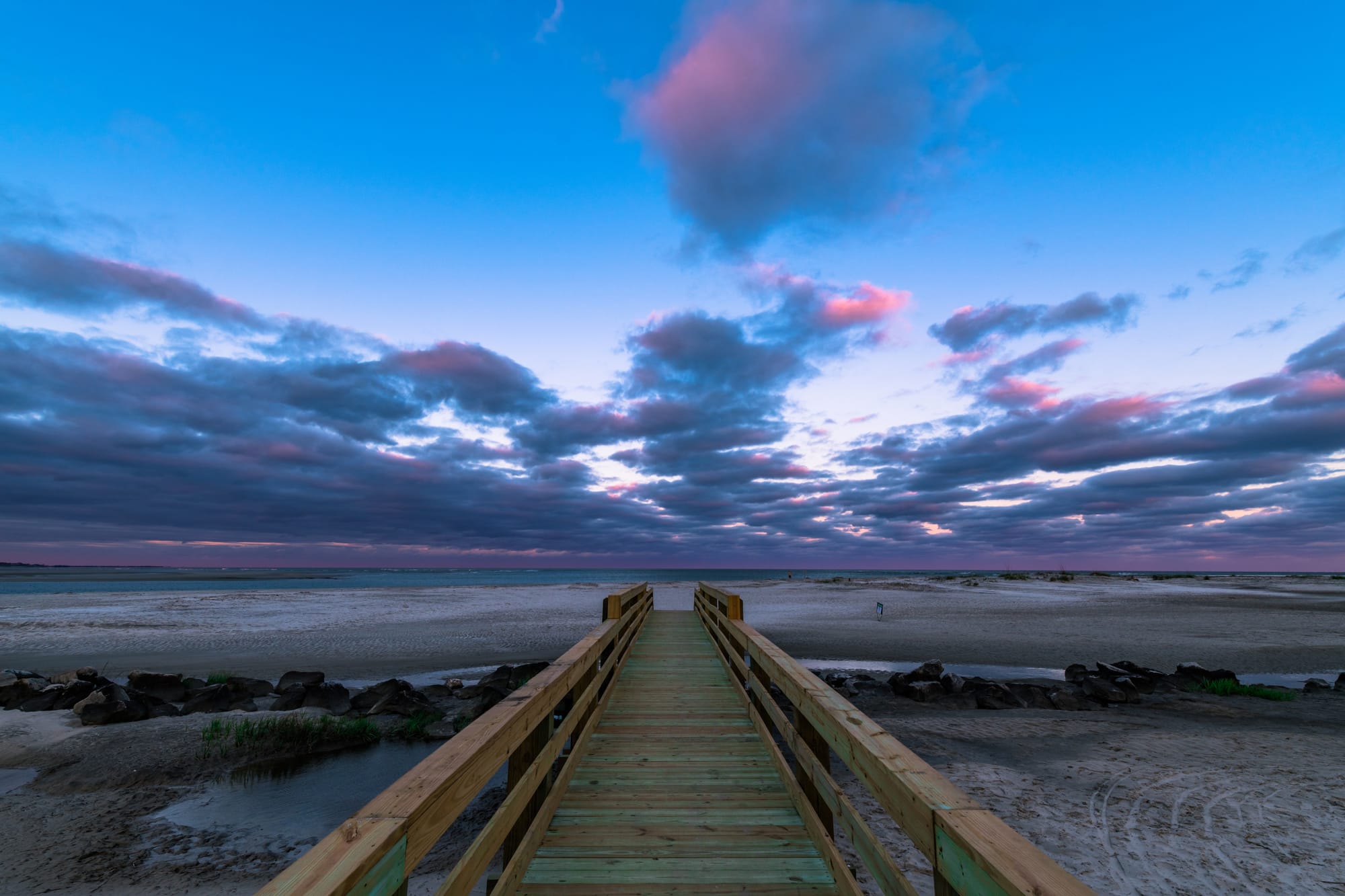 Wooden boardwalk leading to a wide sandy beach under a dramatic sky with pink and purple clouds during twilight.