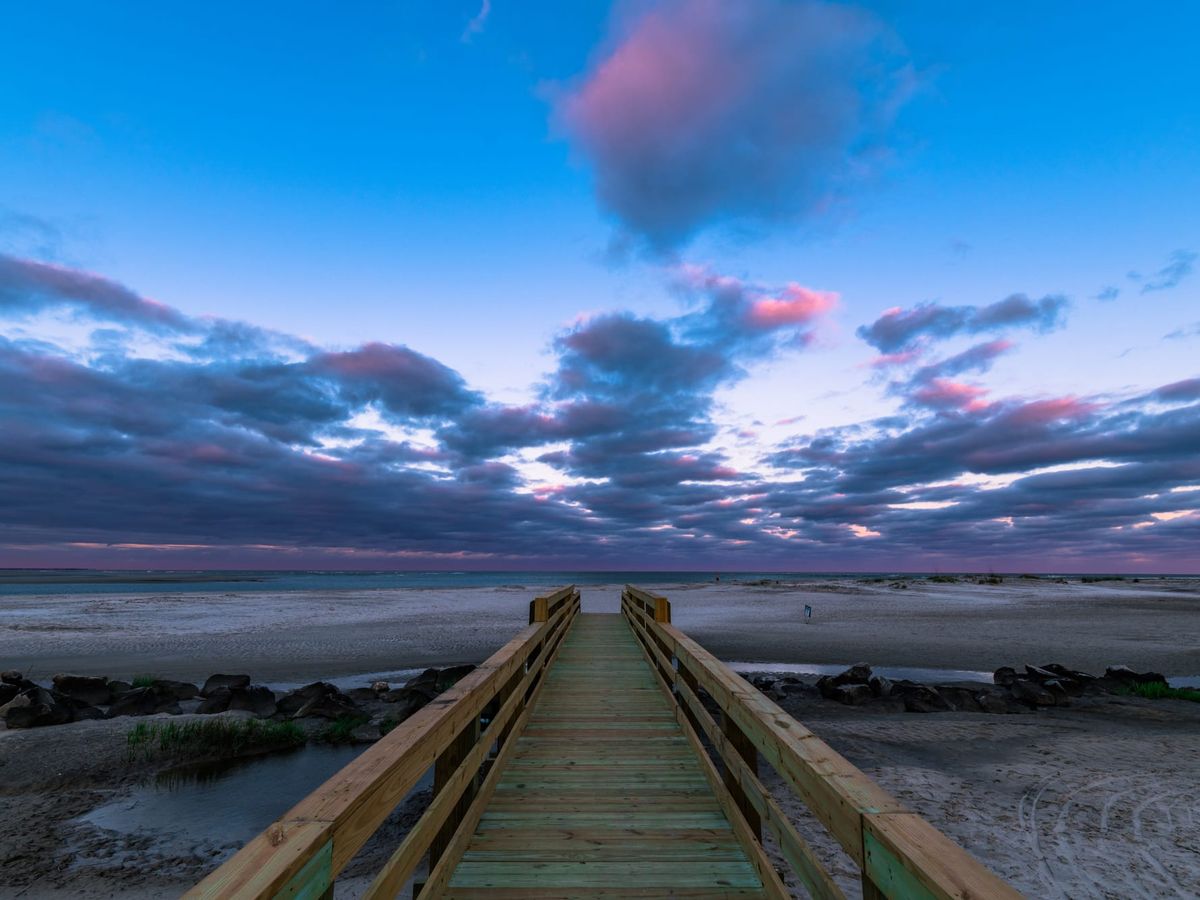 Wooden boardwalk leading to a wide sandy beach under a dramatic sky with pink and purple clouds during twilight.