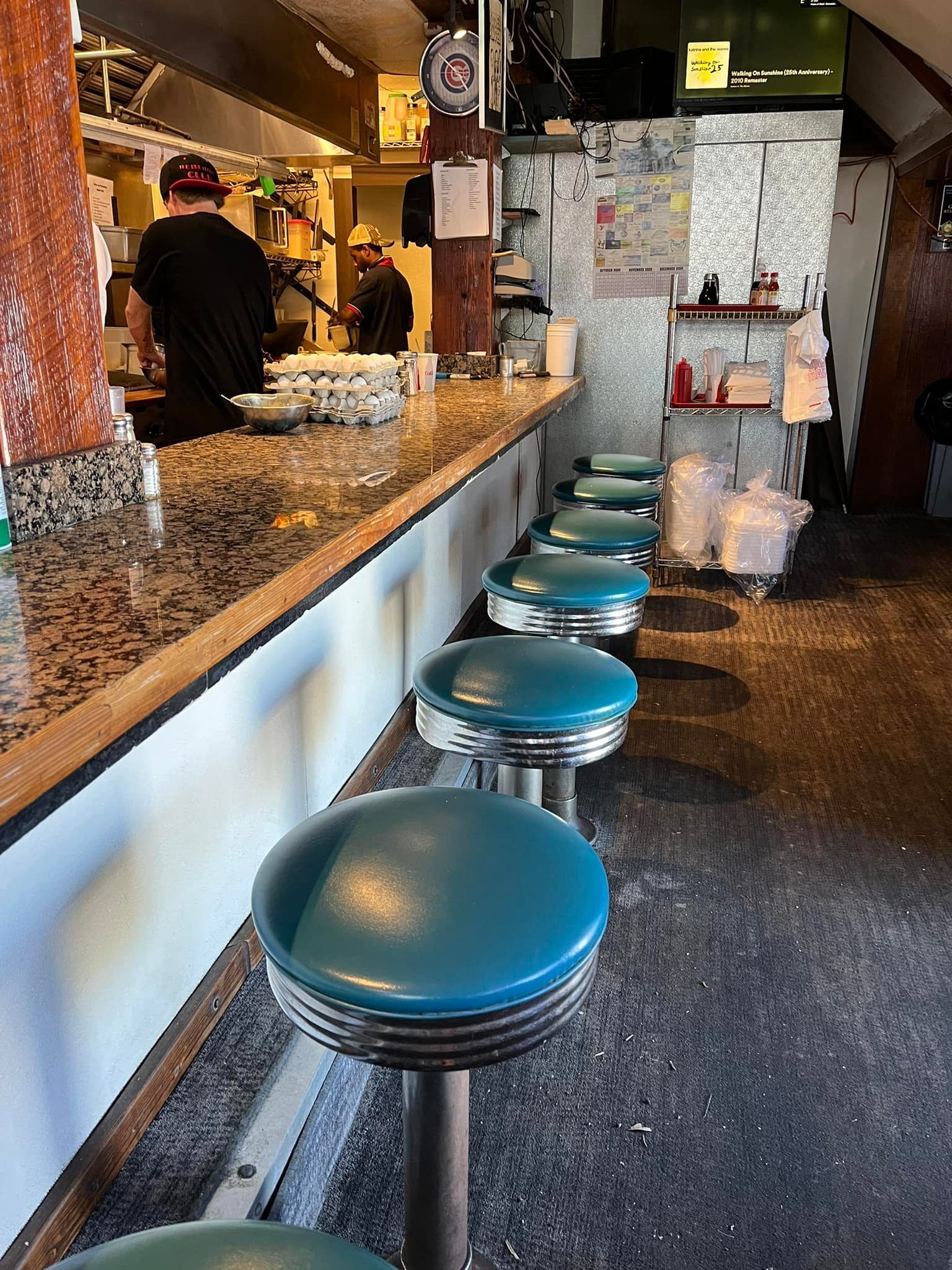 A row of teal vinyl-topped swivel stools lines a granite counter inside the cozy, retro Breakfast Club diner, with two cooks working the grill and cartons of fresh eggs stacked on the counter behind. Warm interior light and vintage decor give the narrow space a classic diner feel. 