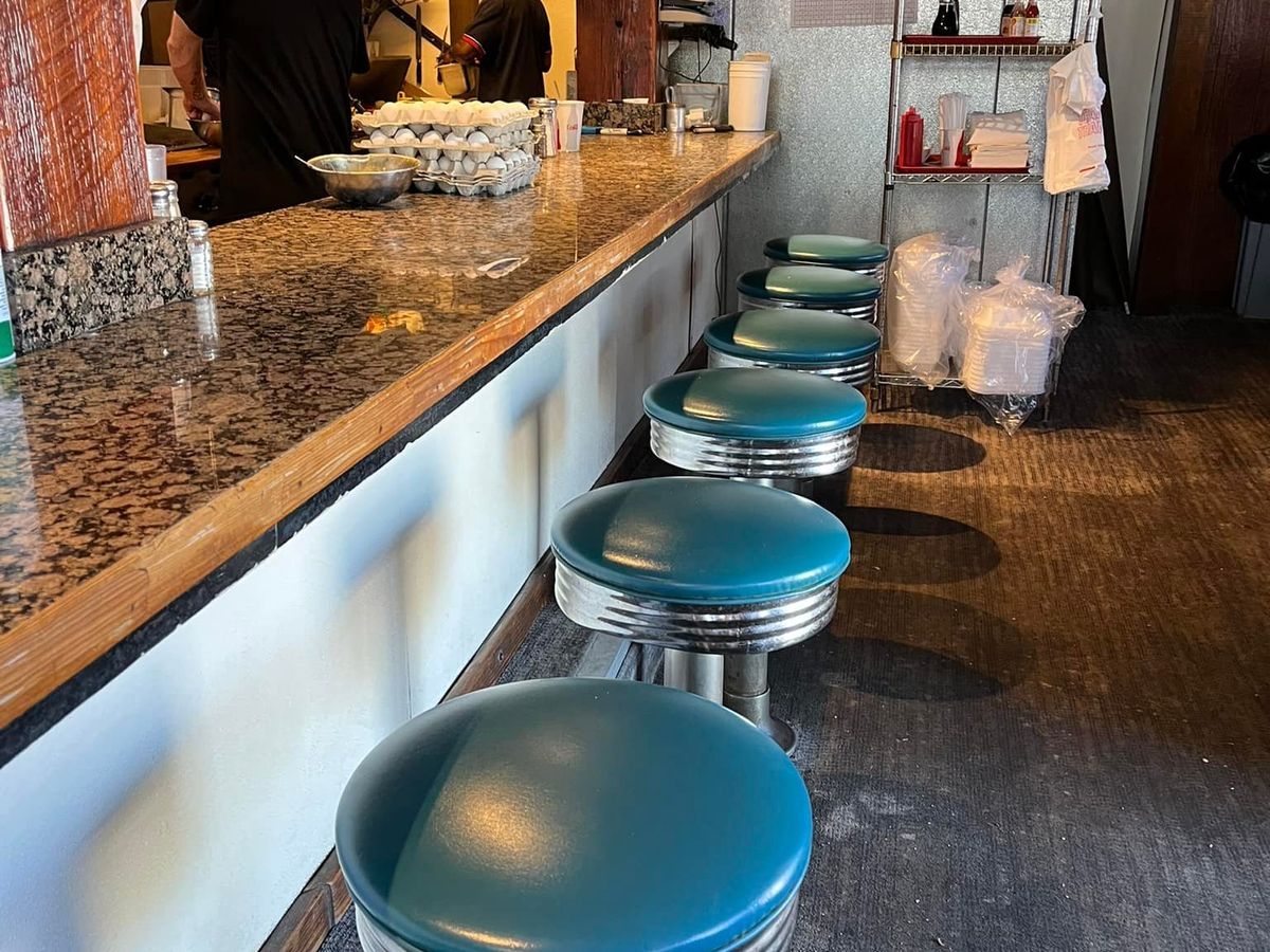 A row of teal vinyl-topped swivel stools lines a granite counter inside the cozy, retro Breakfast Club diner, with two cooks working the grill and cartons of fresh eggs stacked on the counter behind. Warm interior light and vintage decor give the narrow space a classic diner feel.