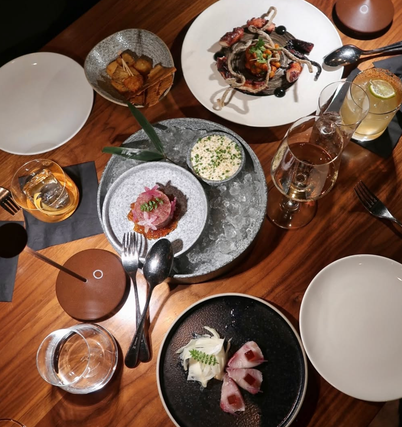 Overhead view of an upscale dining spread featuring artfully plated seafood and small plates, craft cocktails, and wine glasses arranged on a polished wooden table.