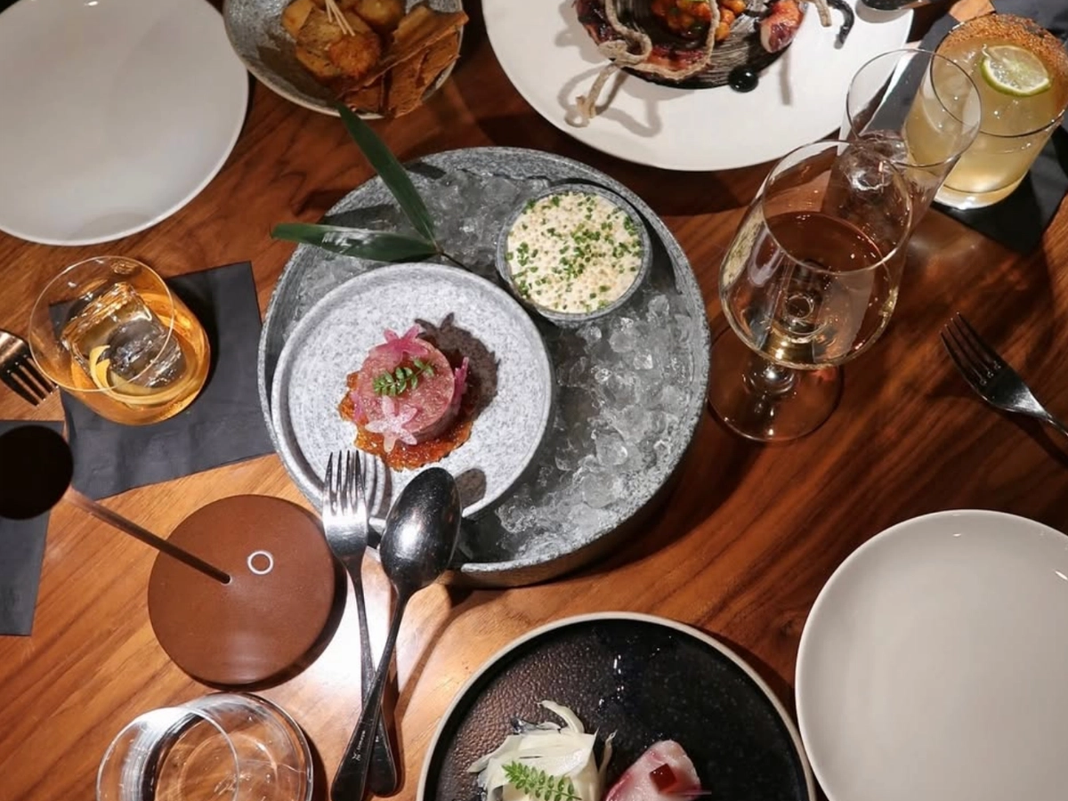 Overhead view of an upscale dining spread featuring artfully plated seafood and small plates, craft cocktails, and wine glasses arranged on a polished wooden table.
