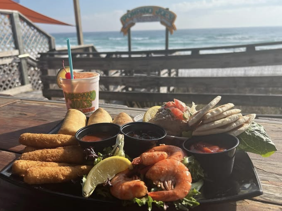 Seafood appetizer platter with shrimp, fish sticks, pita bread, dips, and a frozen cocktail from Pompano Joe’s, served on a wooden deck overlooking the ocean.
