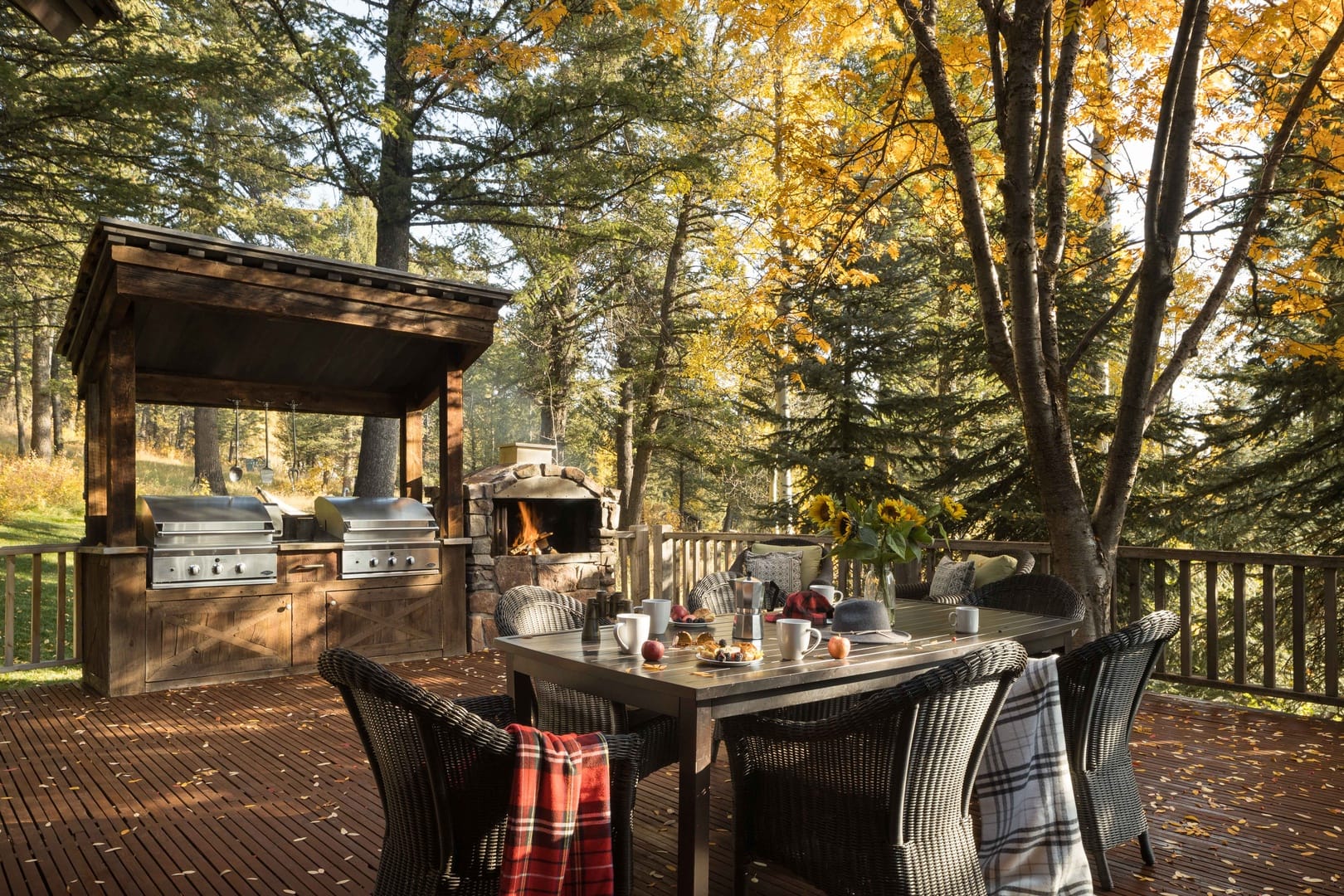 An inviting outdoor deck features a dining table set with coffee and pastries, cozy plaid blankets draped over wicker chairs, and sunflowers as a centerpiece, with twin grills and a stone fireplace surrounded by autumn trees in the background.