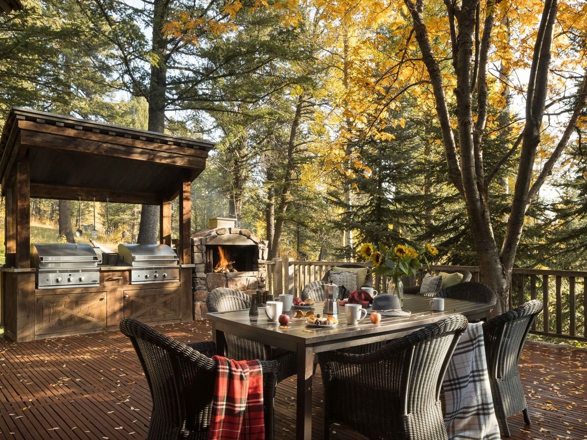 An inviting outdoor deck features a dining table set with coffee and pastries, cozy plaid blankets draped over wicker chairs, and sunflowers as a centerpiece, with twin grills and a stone fireplace surrounded by autumn trees in the background.