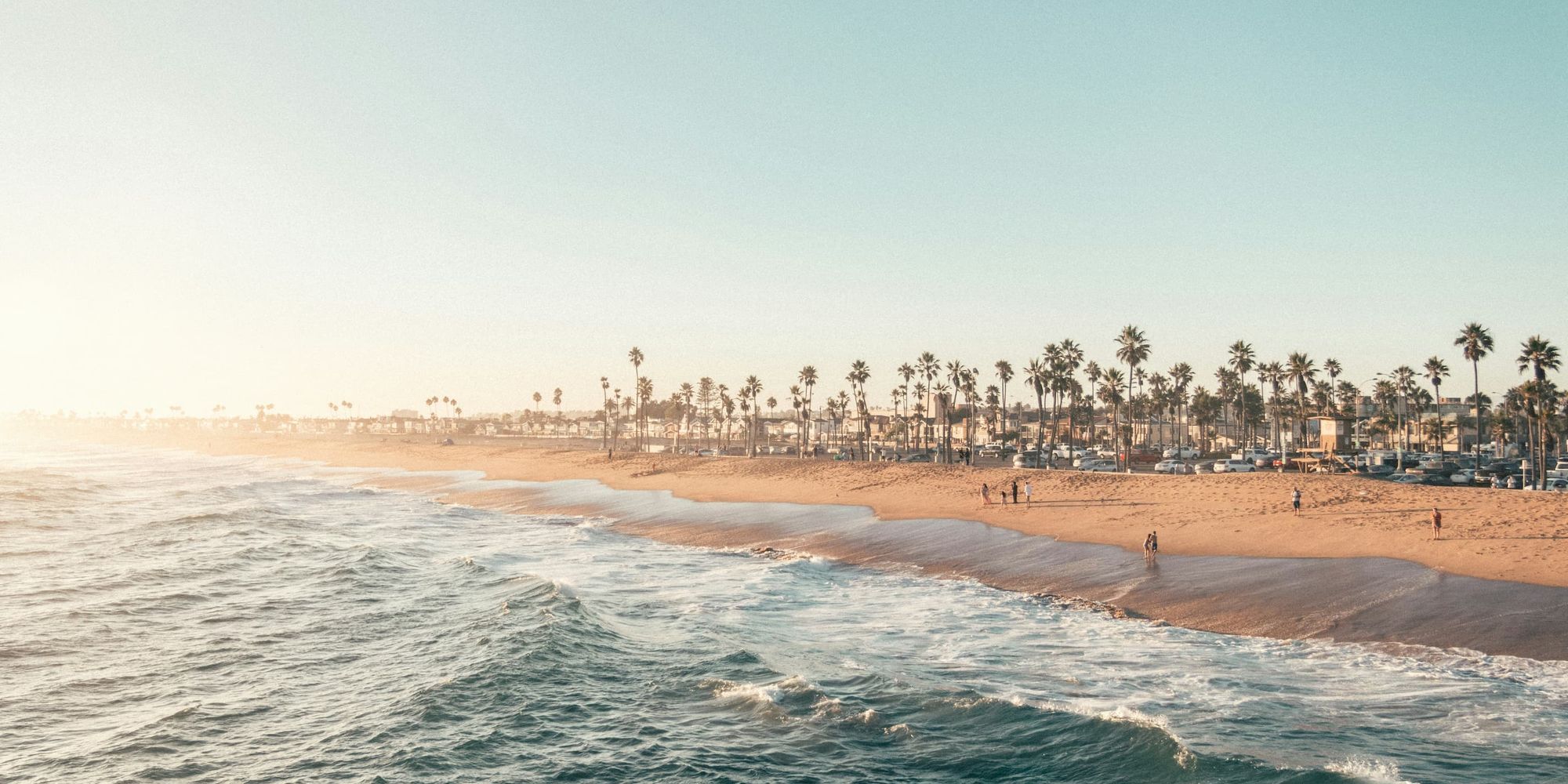 A soft sunset over Newport Beach with golden sand, palm trees, and gentle ocean waves. The scene captures the relaxed, coastal charm that draws travelers to this California favorite year-round.
