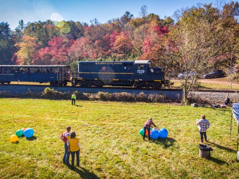 A scenic train ride winds through the Blue Ridge Mountains, passing vibrant fall foliage. Families wave from the grassy field as the historic train rolls by. The colorful autumn leaves and open countryside capture the charm of mountain rail travel.
