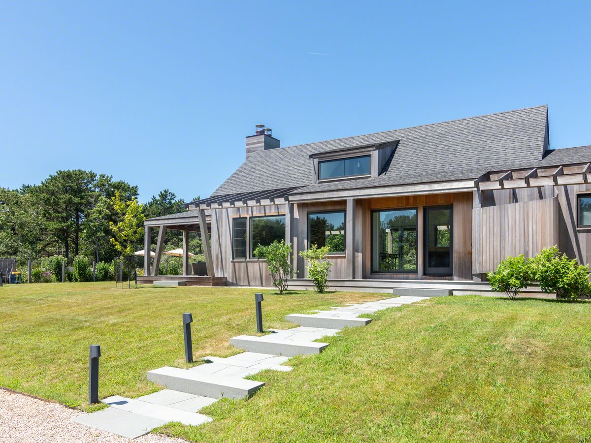 A modern wood-clad home with clean lines and a shaded porch sits on a manicured lawn, bordered by trees under a clear blue sky.