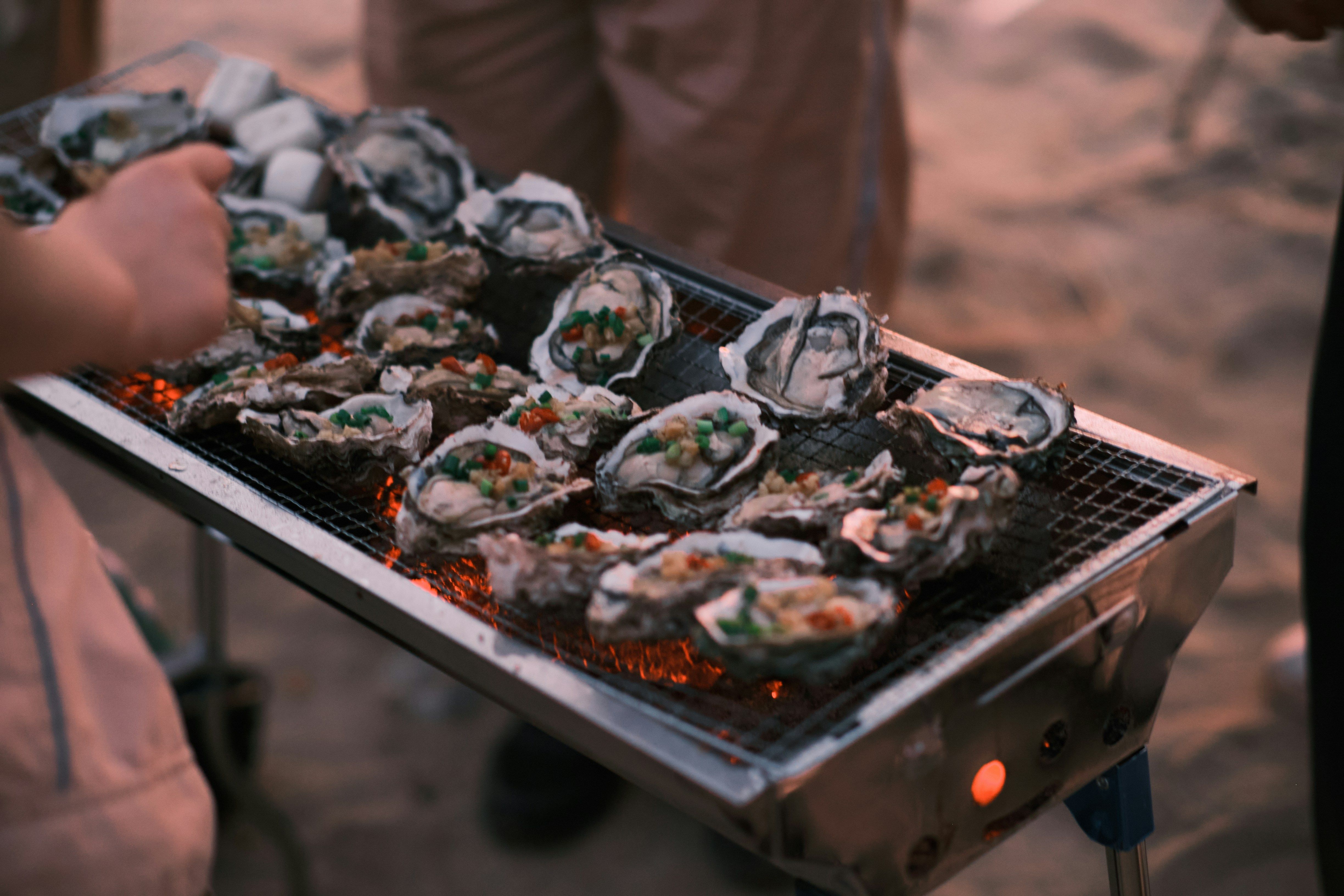 Fresh oysters grilling over open coals on a metal barbecue, topped with chopped vegetables, as people cook and gather around on a sandy beach at dusk.