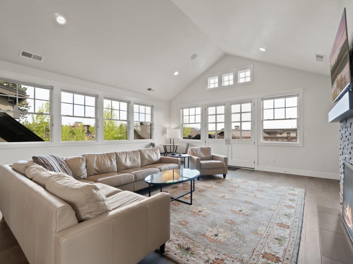 A bright and spacious living room filled with natural light from tall windows, featuring a cozy beige sectional, stone fireplace, and soft patterned rug. The open layout and elegant neutral tones create a warm and inviting retreat for gatherings