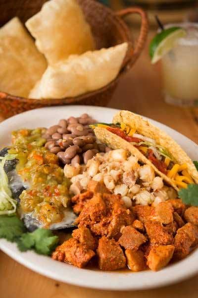 A hearty New Mexican combination plate holds a cheese enchilada smothered in green chile, a crispy taco, seasoned pork adovada, pinto beans, and posole beside a basket of chips and a margarita. The colorful spread showcases classic Southwestern comfort food.