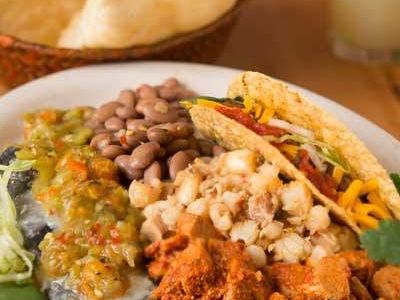 A hearty New Mexican combination plate holds a cheese enchilada smothered in green chile, a crispy taco, seasoned pork adovada, pinto beans, and posole beside a basket of chips and a margarita. The colorful spread showcases classic Southwestern comfort food.