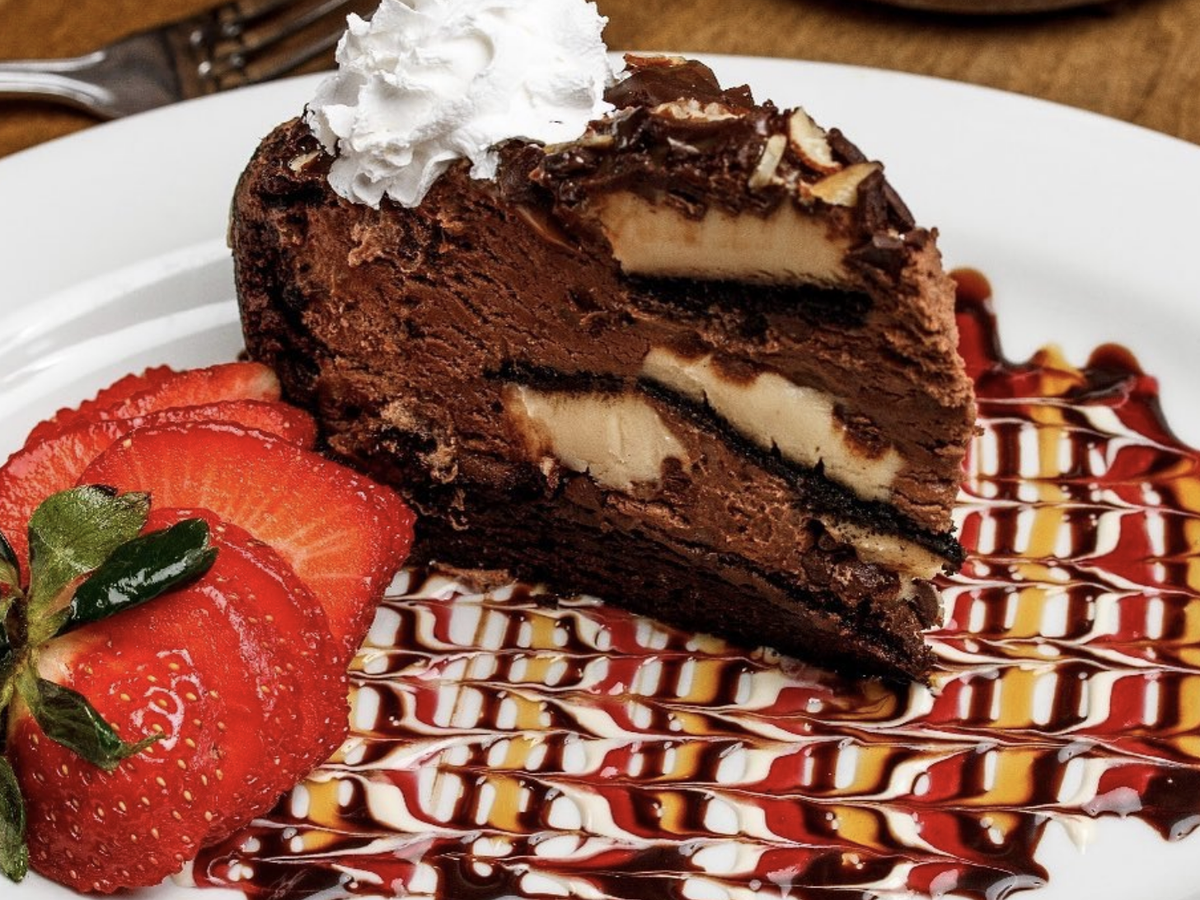 Decadent slice of chocolate cheesecake layered with cream and cookie crust, topped with whipped cream and served with fresh strawberries and drizzled sauces.