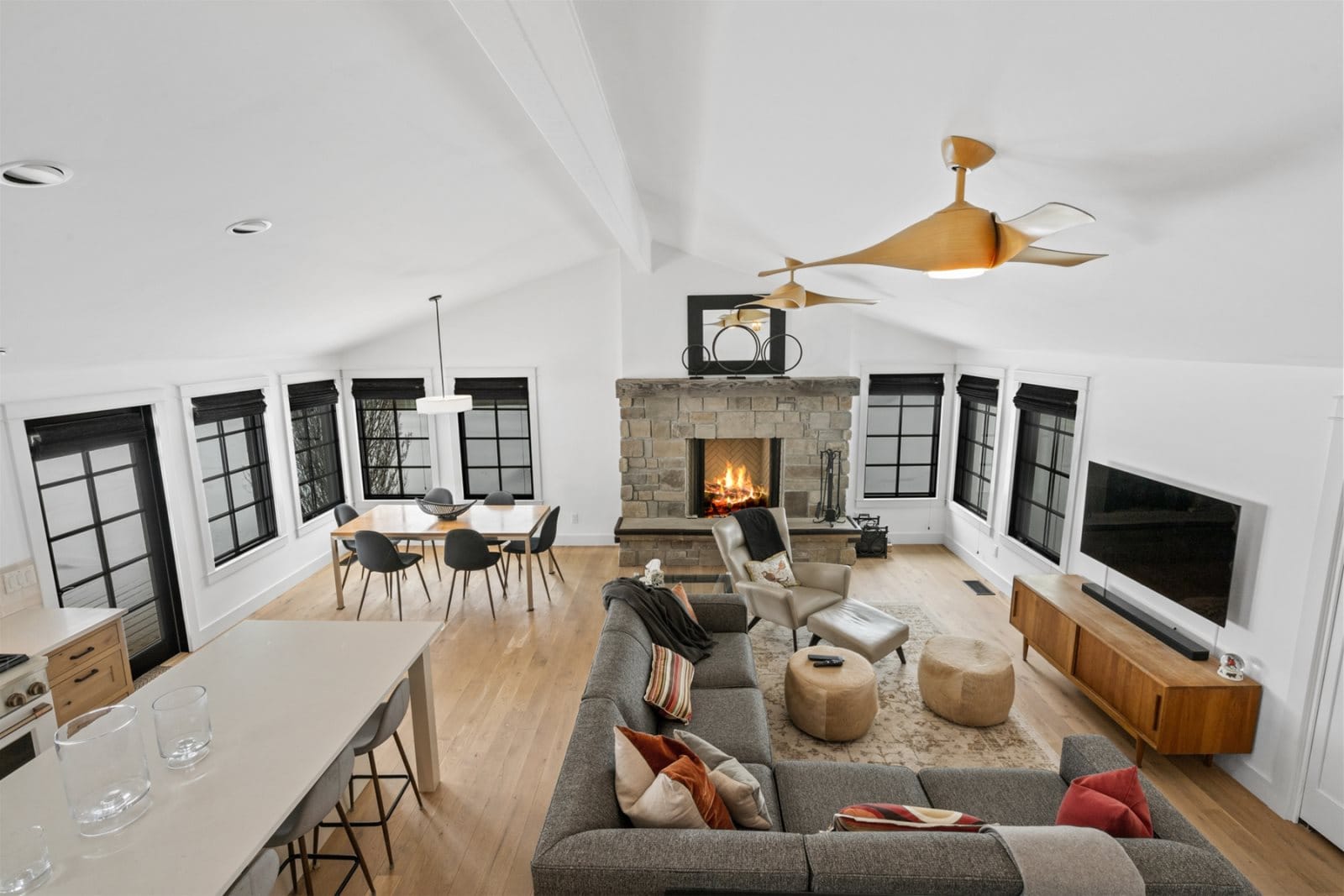 A modern open-concept living and dining area with a stone fireplace, sleek gray sectional sofa, and large black-trimmed windows letting in natural light.