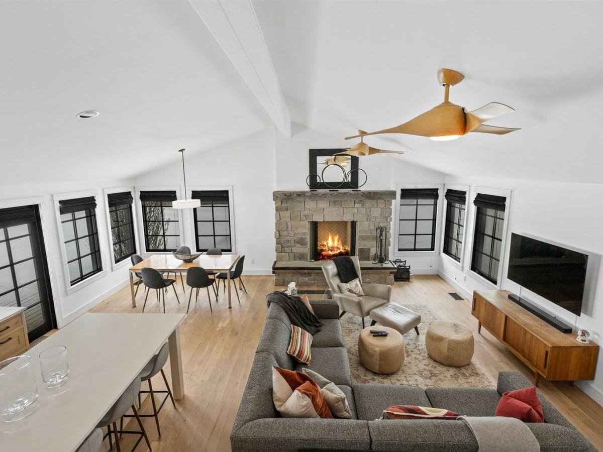 A modern open-concept living and dining area with a stone fireplace, sleek gray sectional sofa, and large black-trimmed windows letting in natural light.