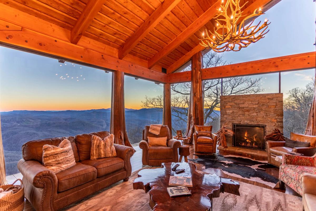 he Flying Eagle's living room at Eagles Nest offers an extraordinary sunset view of the Blue Ridge Mountains through floor-to-ceiling glass walls, framed by soaring timber beams and an antler chandelier overhead. Leather sofas and armchairs gather around a live-edge coffee table and a stacked stone fireplace, creating an elevated mountain lodge atmosphere. 