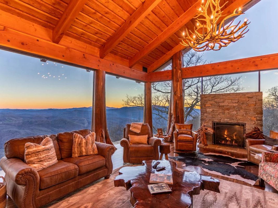 he Flying Eagle's living room at Eagles Nest offers an extraordinary sunset view of the Blue Ridge Mountains through floor-to-ceiling glass walls, framed by soaring timber beams and an antler chandelier overhead. Leather sofas and armchairs gather around a live-edge coffee table and a stacked stone fireplace, creating an elevated mountain lodge atmosphere.