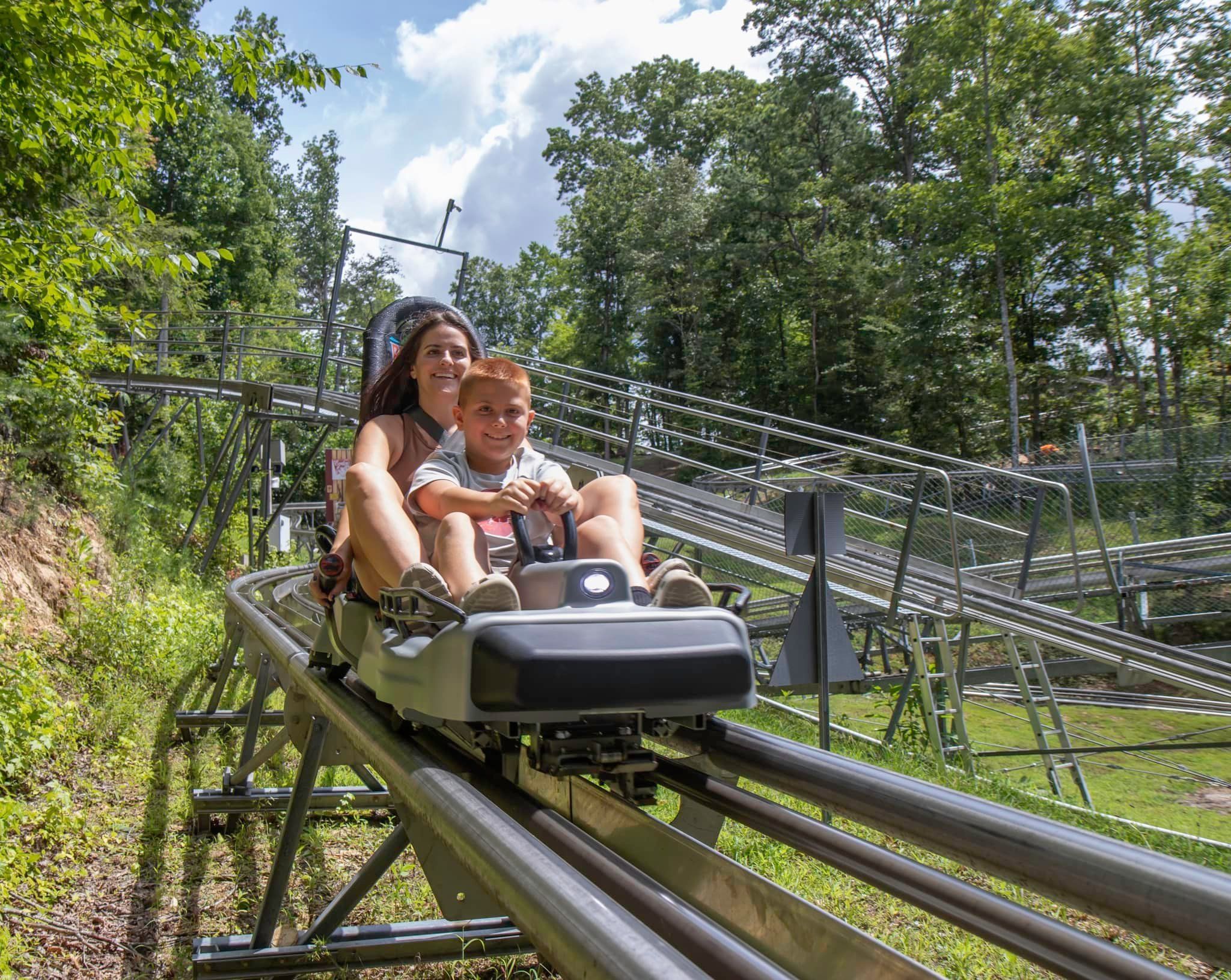 A mom and her young son enjoy an exciting ride down the Rocky Top Mountain Coaster surrounded by lush green trees. The coaster’s metal tracks curve through the Smoky Mountain forest for a fun family adventure.
