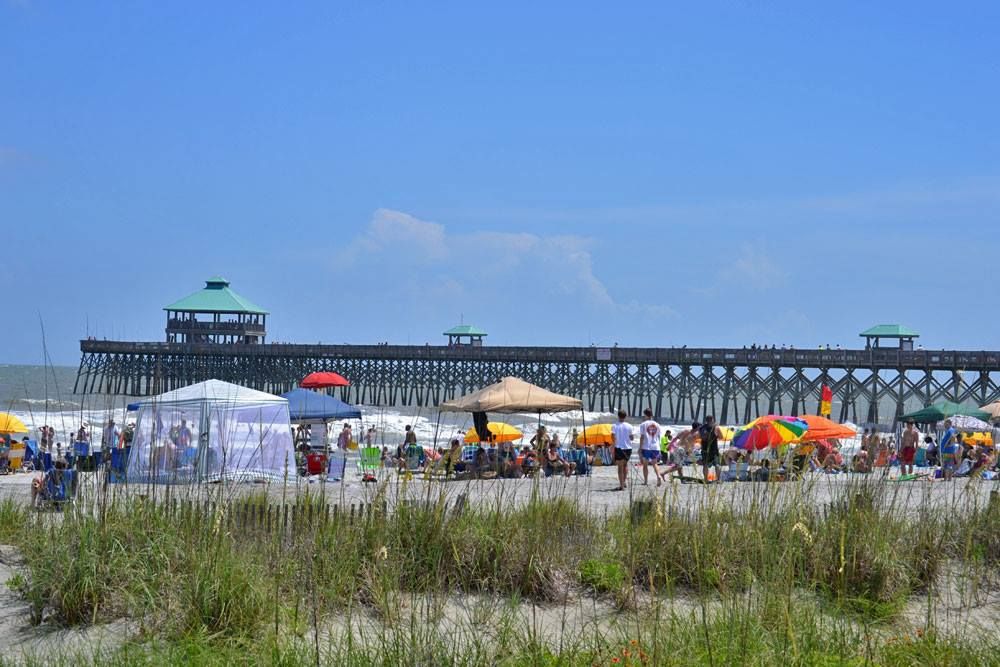A wide sandy beach at Folly Beach with surfers laying on boards and getting ready for a lesson. The open shoreline and calm waves make this a popular spot for beginners learning to surf in Charleston. It’s a laid-back beach scene perfect for warm, sunny days.