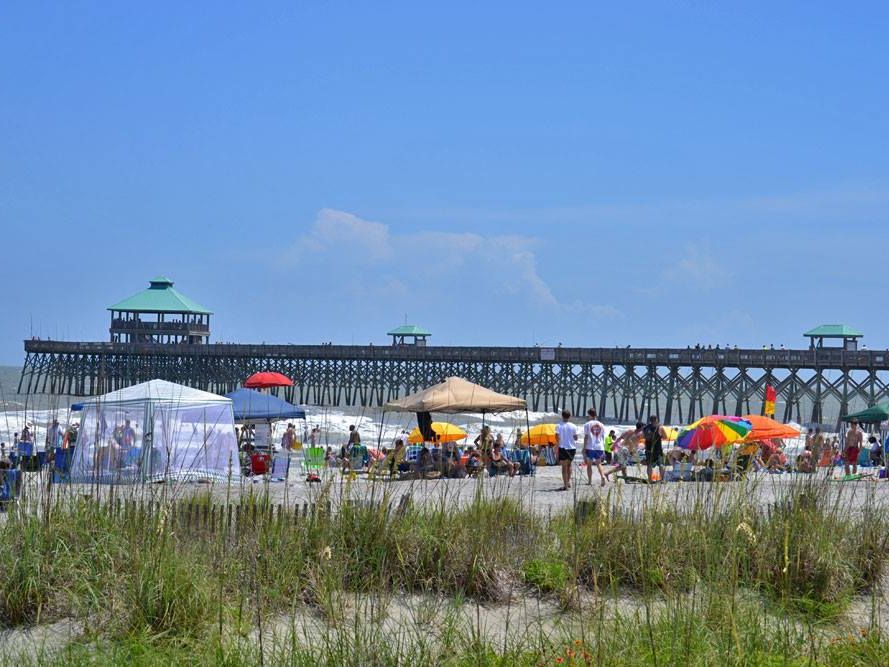 A wide sandy beach at Folly Beach with surfers laying on boards and getting ready for a lesson. The open shoreline and calm waves make this a popular spot for beginners learning to surf in Charleston. It’s a laid-back beach scene perfect for warm, sunny days.