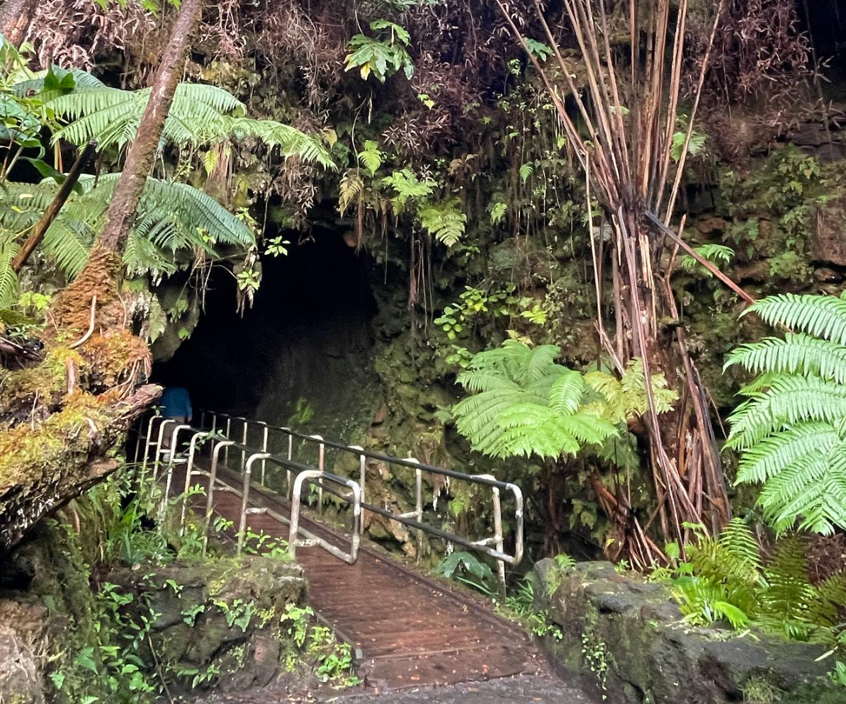 Entrance to the Thurston Lava Tube in Hawai’i Volcanoes National Park, with a wooden walkway and railings leading into the dark tunnel surrounded by lush green ferns and rainforest.