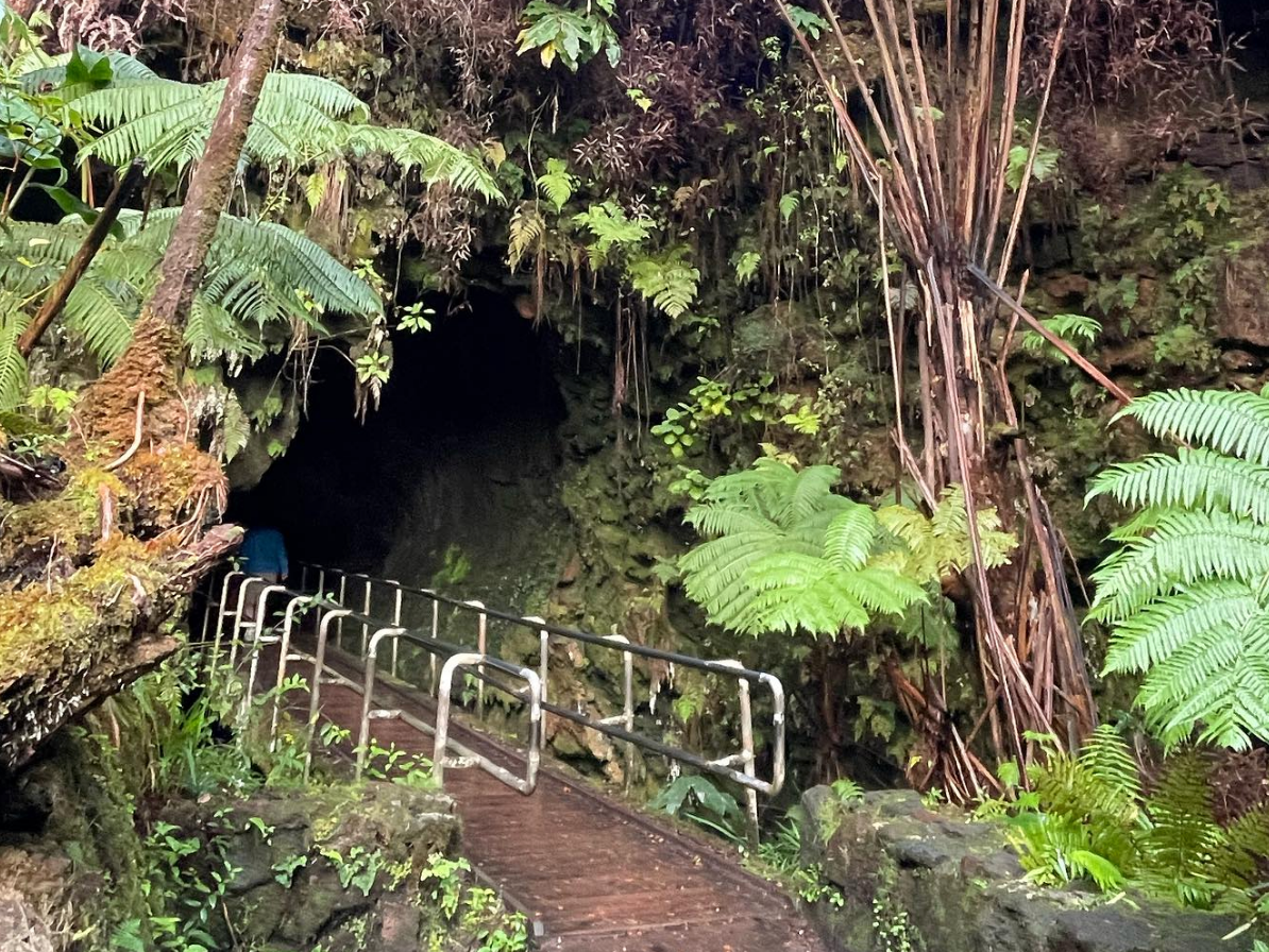 Entrance to the Thurston Lava Tube in Hawai’i Volcanoes National Park, with a wooden walkway and railings leading into the dark tunnel surrounded by lush green ferns and rainforest.