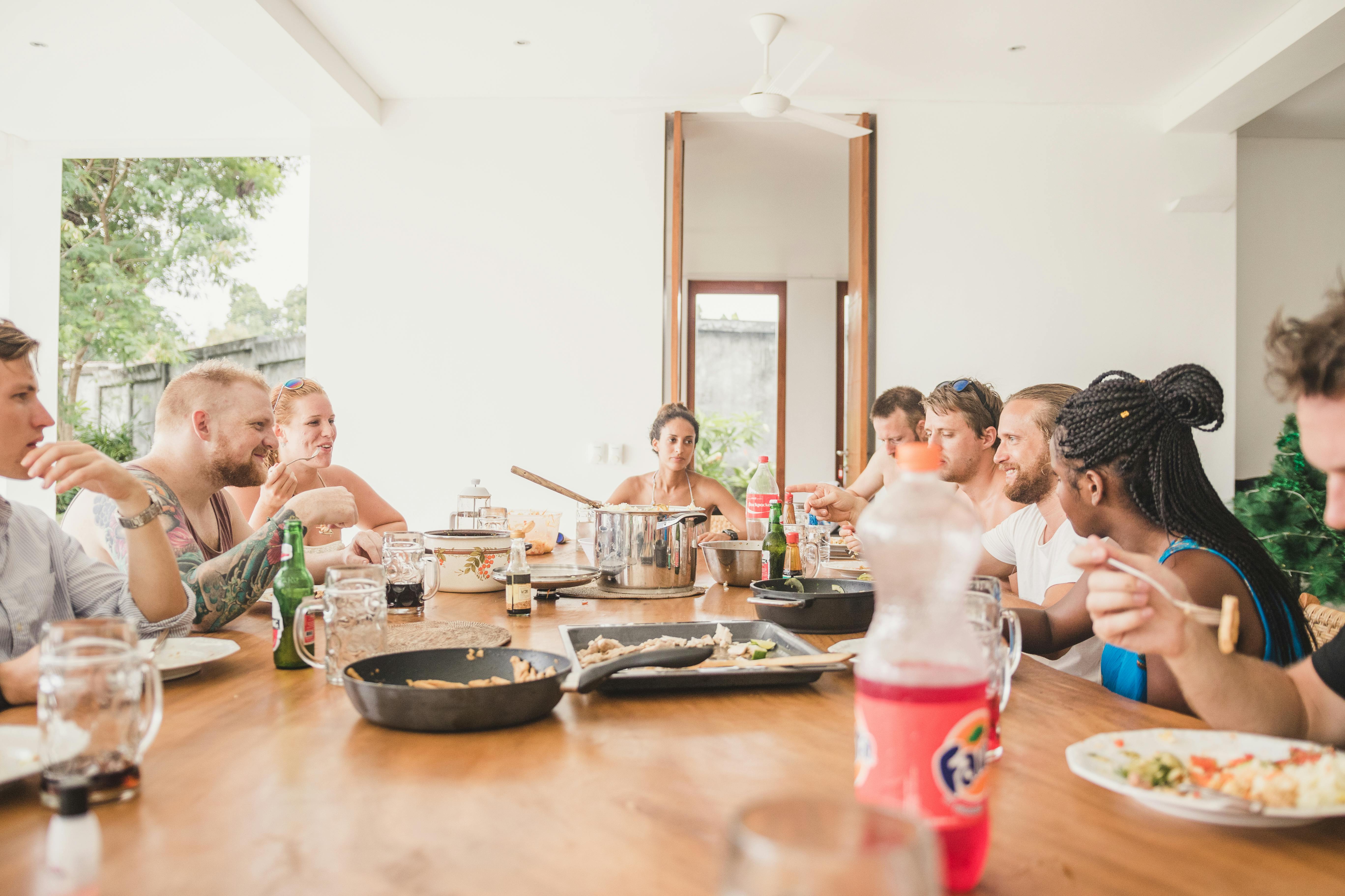 A group of friends enjoying a shared meal at a large wooden dining table filled with plates, drinks, and serving dishes. The warm, casual setting captures the joy of togetherness and shared moments during a vacation stay.