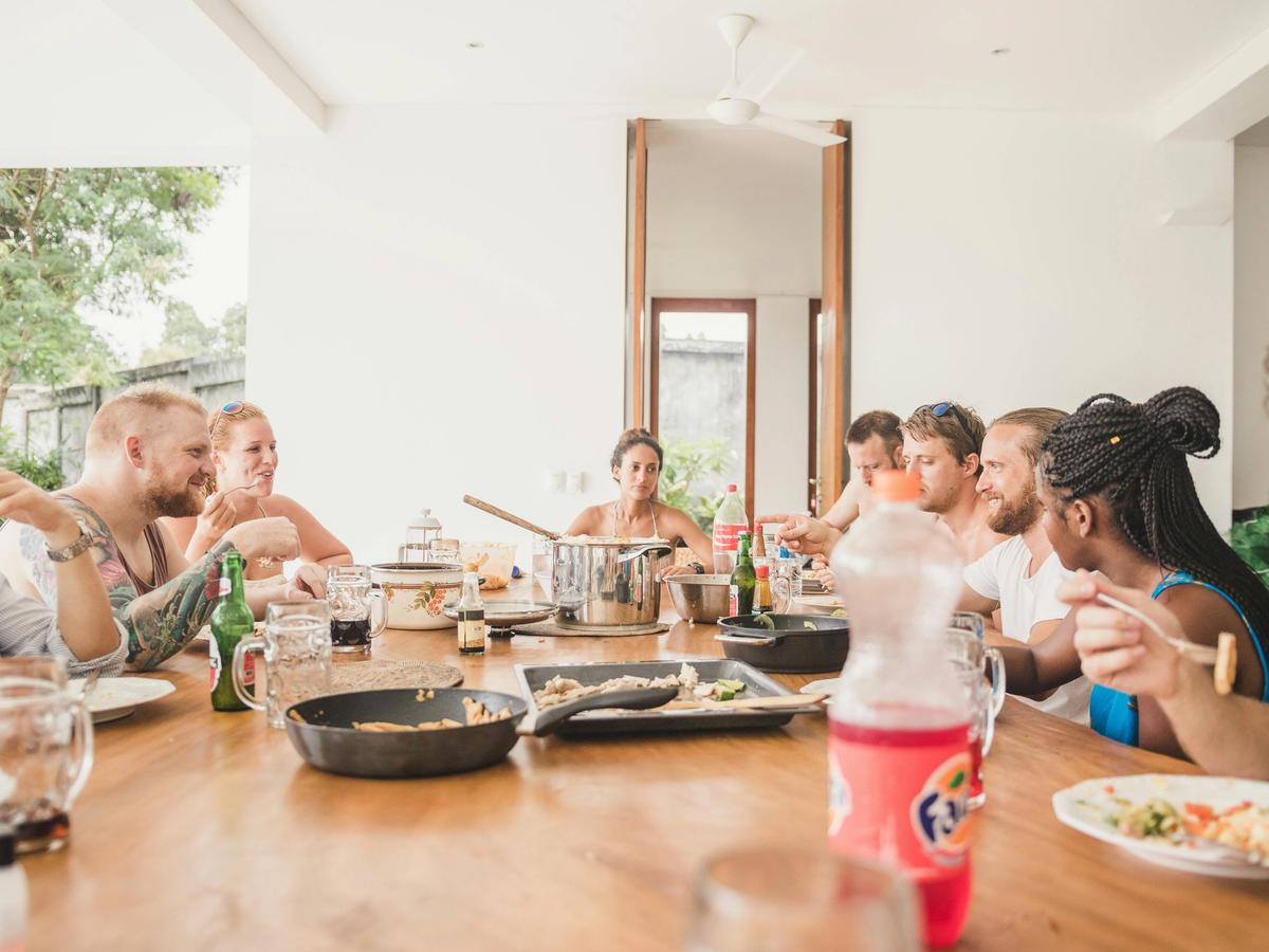 A group of friends enjoying a shared meal at a large wooden dining table filled with plates, drinks, and serving dishes. The warm, casual setting captures the joy of togetherness and shared moments during a vacation stay.