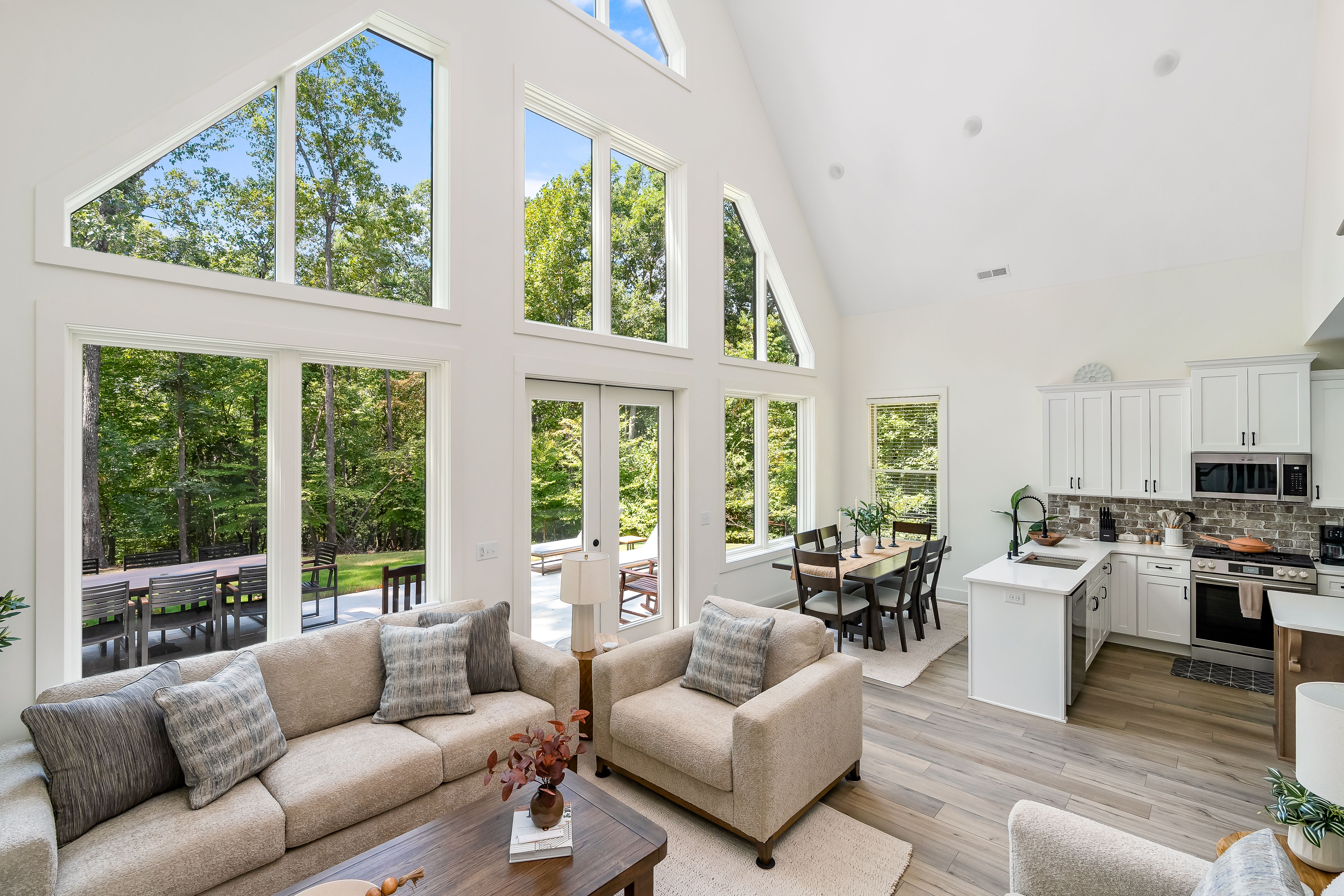 Modern open-concept living room and kitchen with vaulted ceilings, large floor-to-ceiling windows, neutral-toned sofa and chairs, dining area, and white cabinetry with stainless steel appliances.